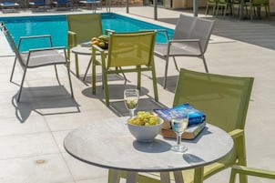 Zona junto a la piscina con una mesa, sillas, un cuenco de aceitunas, una copa de vino y un libro.
