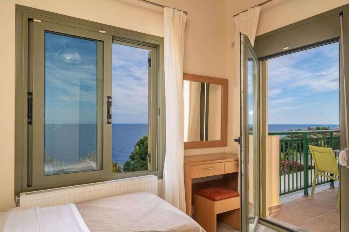 Un dormitori amb llit, escriptori de fusta, mirall i un balcó amb vista al mar.