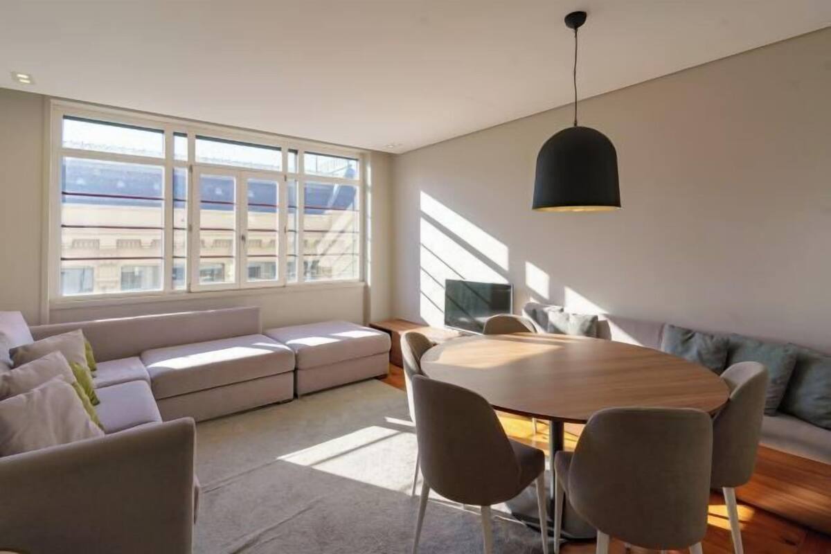 A modern living room with a sofa, a dining table, and chairs. There is a large window providing natural light and a view of buildings outside.