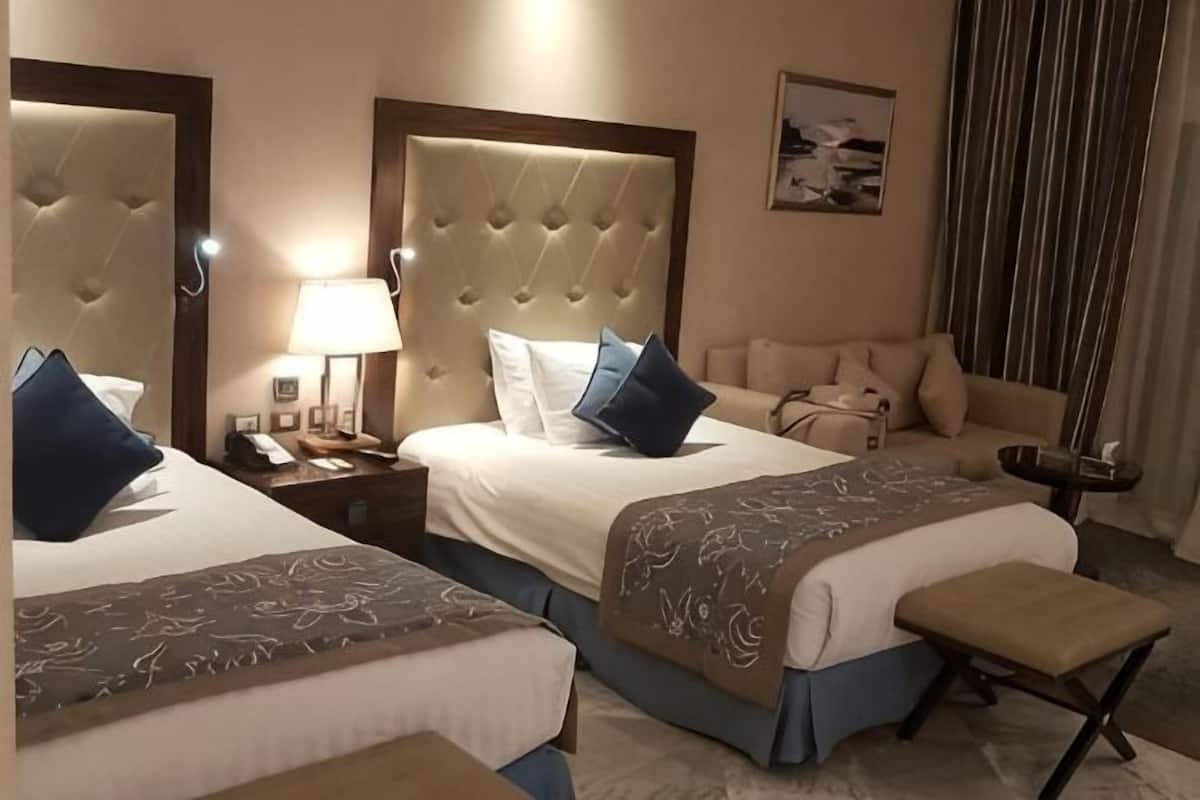 A hotel room with two beds, a sofa, a nightstand, a lamp, and a painting on the wall.
