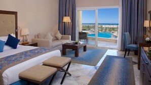 A hotel room with a bed, a sofa, a coffee table, a desk, and a view of a pool and beach. - Ghazala Luxury Collection Alamein (Qesm Ad Dabaah)