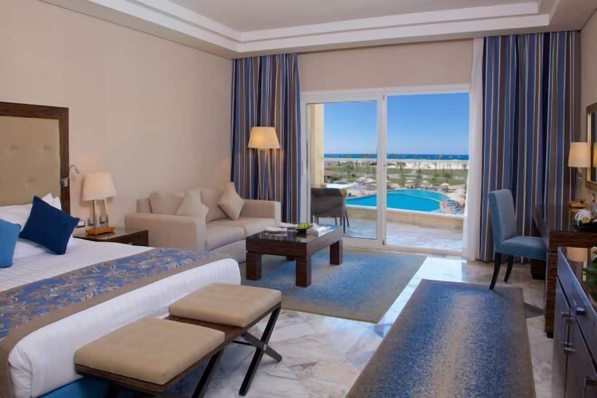 A hotel room with a bed, a sofa, a coffee table, a desk, and a view of a pool and beach.