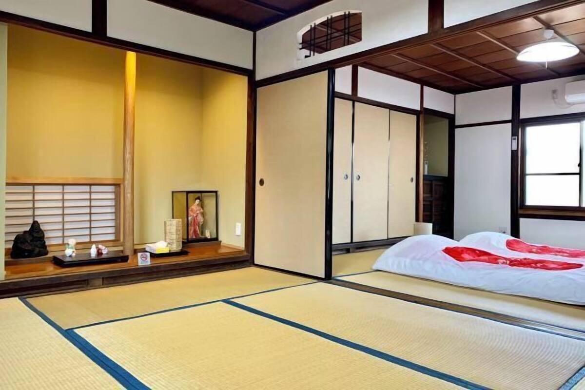 A traditional Japanese room with tatami flooring, a shoji screen, a bed with a red and white bedding, and a small shrine with offerings.