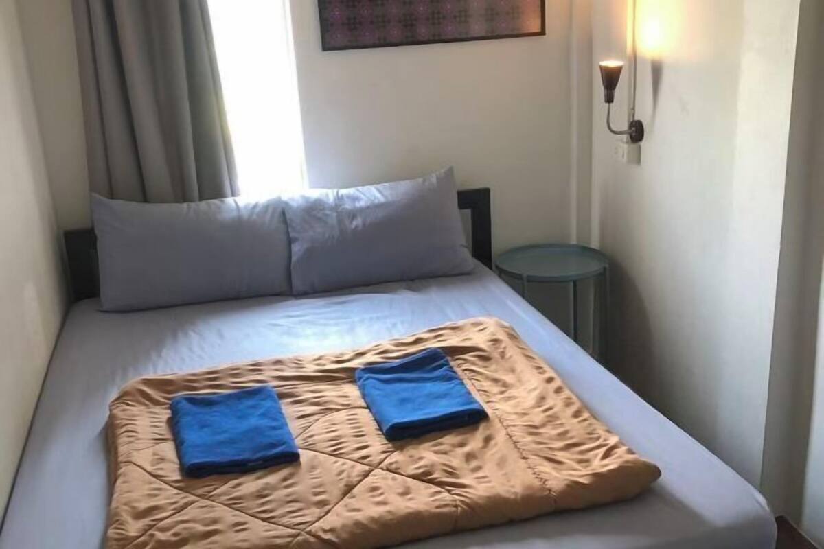 A bed with a beige quilt, two blue towels, a gray pillow, a small round table, and a wall-mounted light fixture.