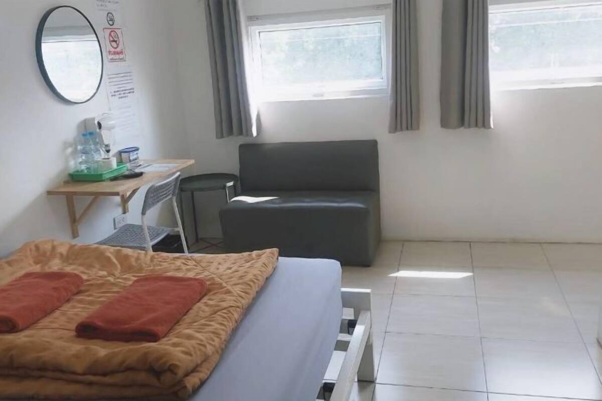 A hotel room with a bed, a desk, a chair, a grey sofa, a window with curtains, and a wall-mounted mirror.
