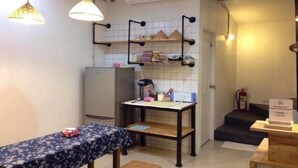 A compact kitchen with a refrigerator, a table with a floral tablecloth, and hanging pendant lights. - Dhub Hostel Donmueng (Bangkok)