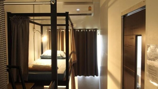 A bunk bed room with a wooden frame, a desk, and a chair. There is a window with curtains, an air conditioning unit, and a door with a poster. - Dhub Hostel Donmueng (Bangkok)
