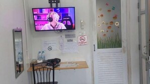 A room with a television mounted on the wall, a desk with a chair, a door with a flower decal, and a no-smoking sign. - Dhub Hostel Donmueng (Bangkok)
