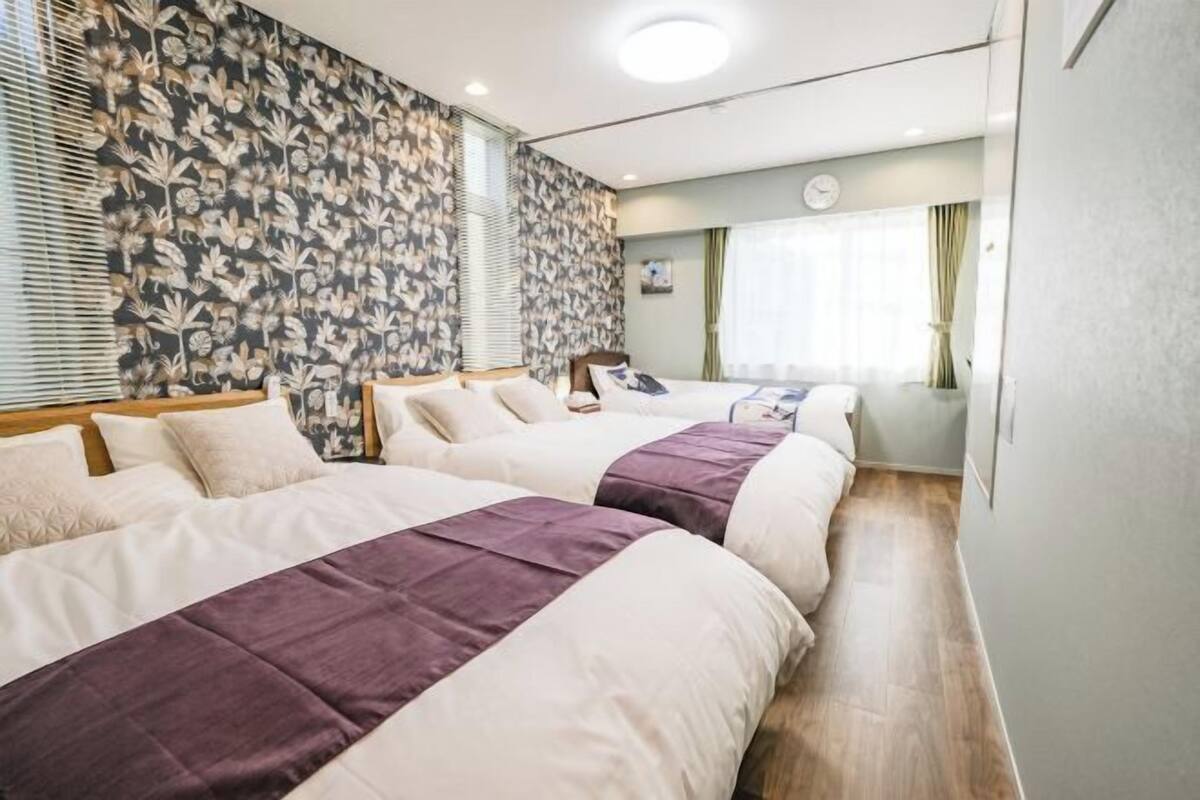 Een hotelkamer met twee bedden, een groot raam met gordijnen, een klok aan de muur en een behang met bloemenmotief.
