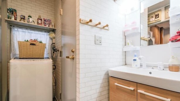 Un baño con pared de ladrillo blanco, un lavabo blanco, armarios de madera, un espejo y una cesta sobre el radiador.