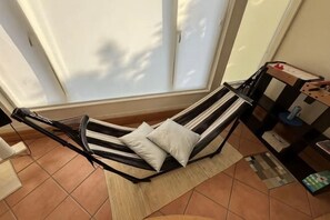 Una silla hamaca con almohadones, piso de baldosas, una ventana con persianas y una mesita con objetos.