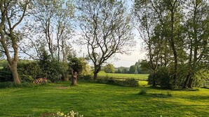 Ein gepflegter Rasen mit einem Kiesweg, der zu einer Sitzgelegenheit führt, umgeben von alten Bäumen und verschiedenen blühenden Pflanzen.