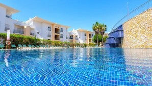 A swimming pool with clear blue water, surrounded by white buildings with balconies, and a stone wall on one side. - BnB Homes Ocean View Apartment with relax Terrace, 2 Swimming pools & Tennis court (Albufeira)