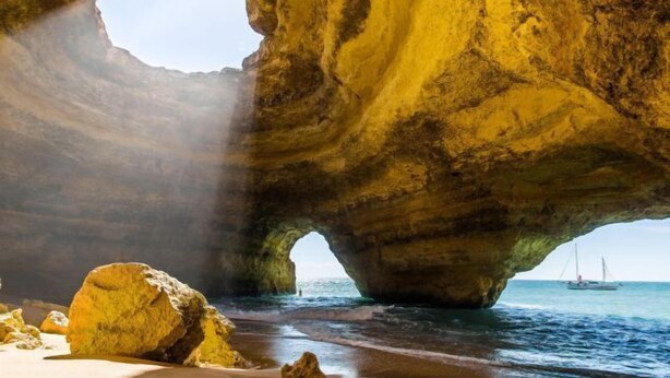A natural rock formation with a cave-like opening, a sandy beach, and a boat in the water. - BnB Homes Ocean View Apartment with relax Terrace, 2 Swimming pools & Tennis court (Albufeira)