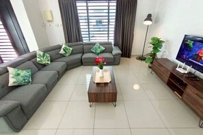 A living room with a grey sectional sofa, a wooden coffee table with a vase of flowers, a TV on a wooden stand, and a floor lamp. - Tropical Livings Meru Ipoh Homestay (Ipoh)