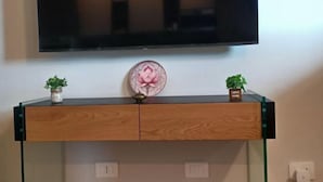 Ein modernes Wohnzimmer mit einem flachbildfernseher an der Wand, einer Holzkommode mit zwei kleinen Topfpflanzen und einem dekorativen Teller mit einem Lotusmuster.