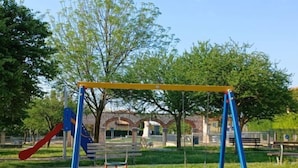 Ein Spielplatz mit Schaukeln und einer Rutsche, umgeben von Bäumen und einem Gebäude im Hintergrund.