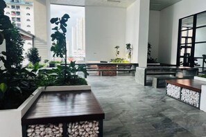 Un espacio interior amplio con un largo banco, plantas en macetas y vistas de edificios altos.