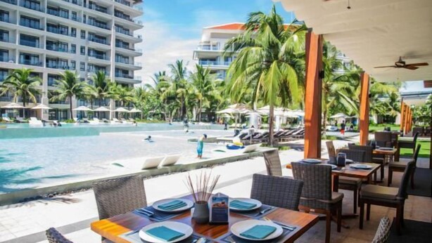 A beachfront restaurant with outdoor seating overlooking a pool and beach, surrounded by palm trees and hotel buildings. - Ocean Villa Da Nang 4BR 5 Beds, Private Pool and Kitchen (Da Nang)