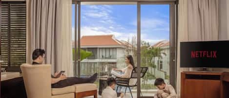 A living room with a large TV, a sofa, a dining table, and a balcony with a view.