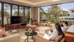 A modern living room with a large TV displaying 'NETFLIX', comfortable seating, and a view of a landscaped area outside. - Ocean Villa Da Nang 4BR 5 Beds, Private Pool and Kitchen (Da Nang)