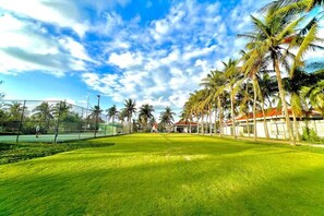 Een verzorgd gazon met een tennisbaan, palmbomen en een gebouw met een rood dak.