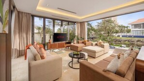A modern living room with a flat-screen TV, comfortable sofas, a coffee table, and large windows offering a view of the outdoors. - Ocean Villa Da Nang 4BR 5 Beds, Private Pool and Kitchen (Da Nang)