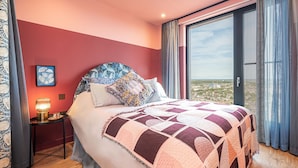 A bedroom with a large bed, a nightstand, a lamp, and a view of the city through a large window. - The Eades Laura Perryman 1 Bed (London)