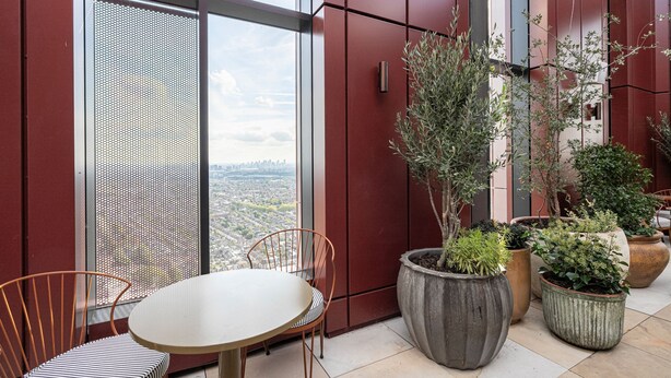 Ein moderner Balkon mit rundem Tisch und Stühlen, großen Kübelpflanzen und Blick auf die Stadt durch ein großes Fenster.