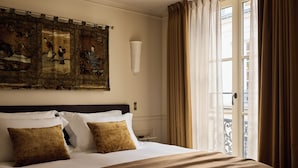 A hotel room with a large bed, a headboard, a wall-mounted artwork, a window with curtains, and a view of the outside. - L'Opale Noire (Paris)