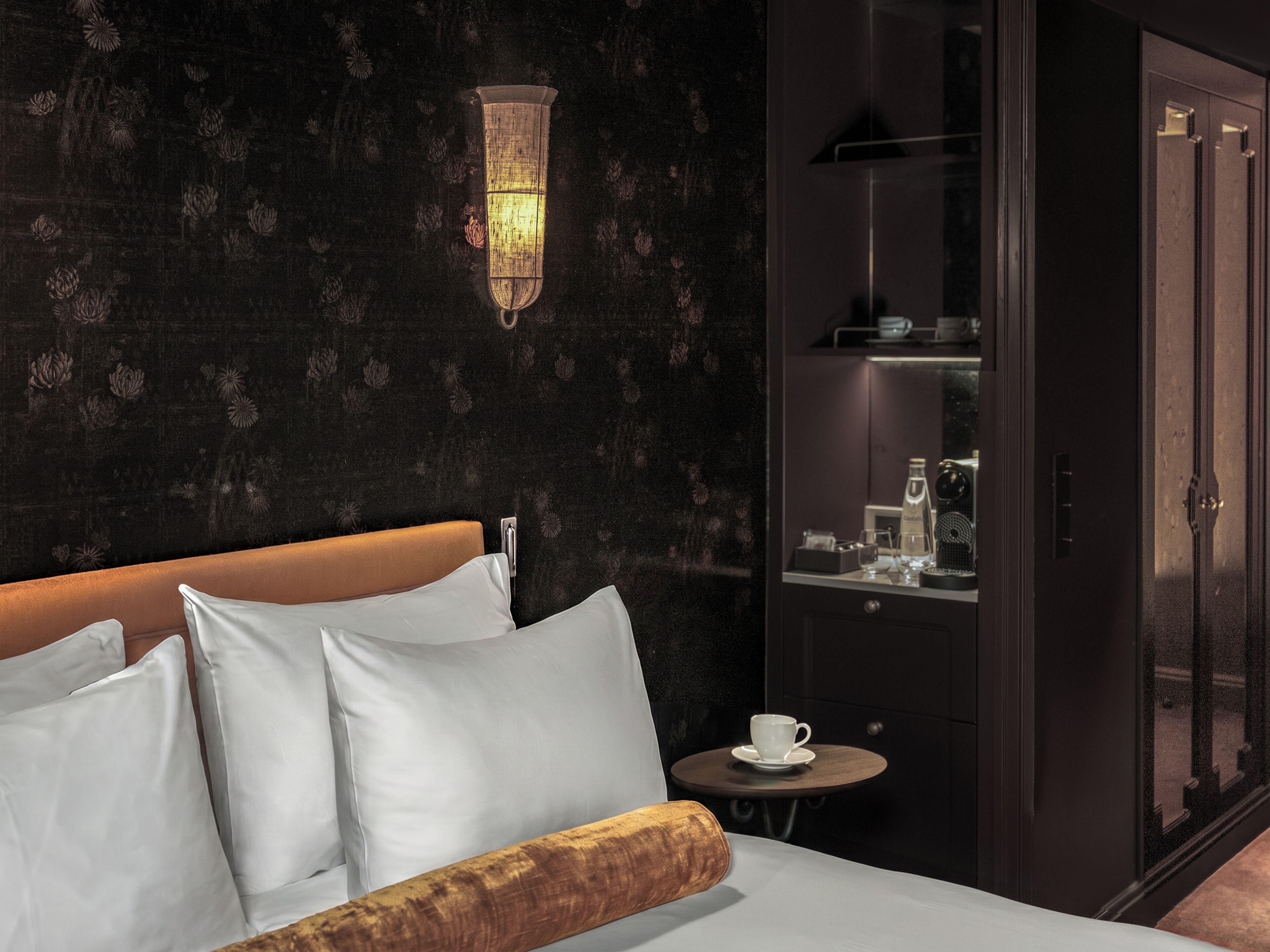 Habitación de hotel con cama, almohadas blancas, cabecero de madera, una pequeña mesa redonda con una taza y una lámpara de pared.