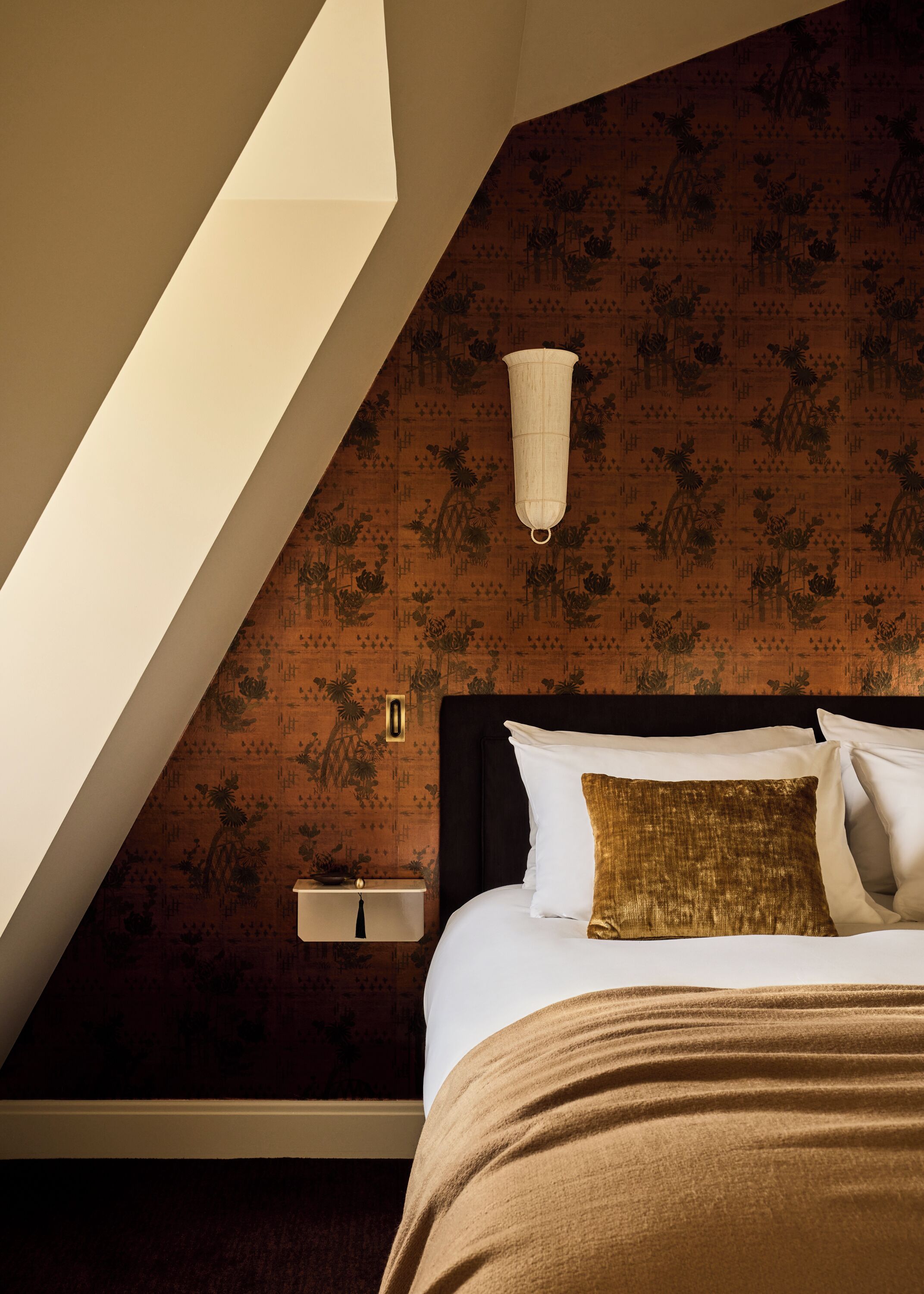 Une chambre avec un plafond en pente, un lit agrémenté d’oreillers blancs et dorés, une table de chevet et un luminaire fixé au mur.