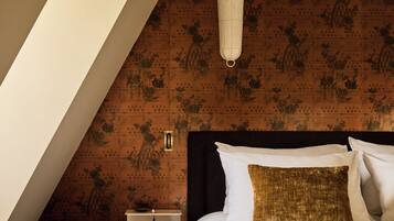 Une chambre avec un plafond en pente, un lit agrémenté d’oreillers blancs et dorés, une table de chevet et un luminaire fixé au mur.