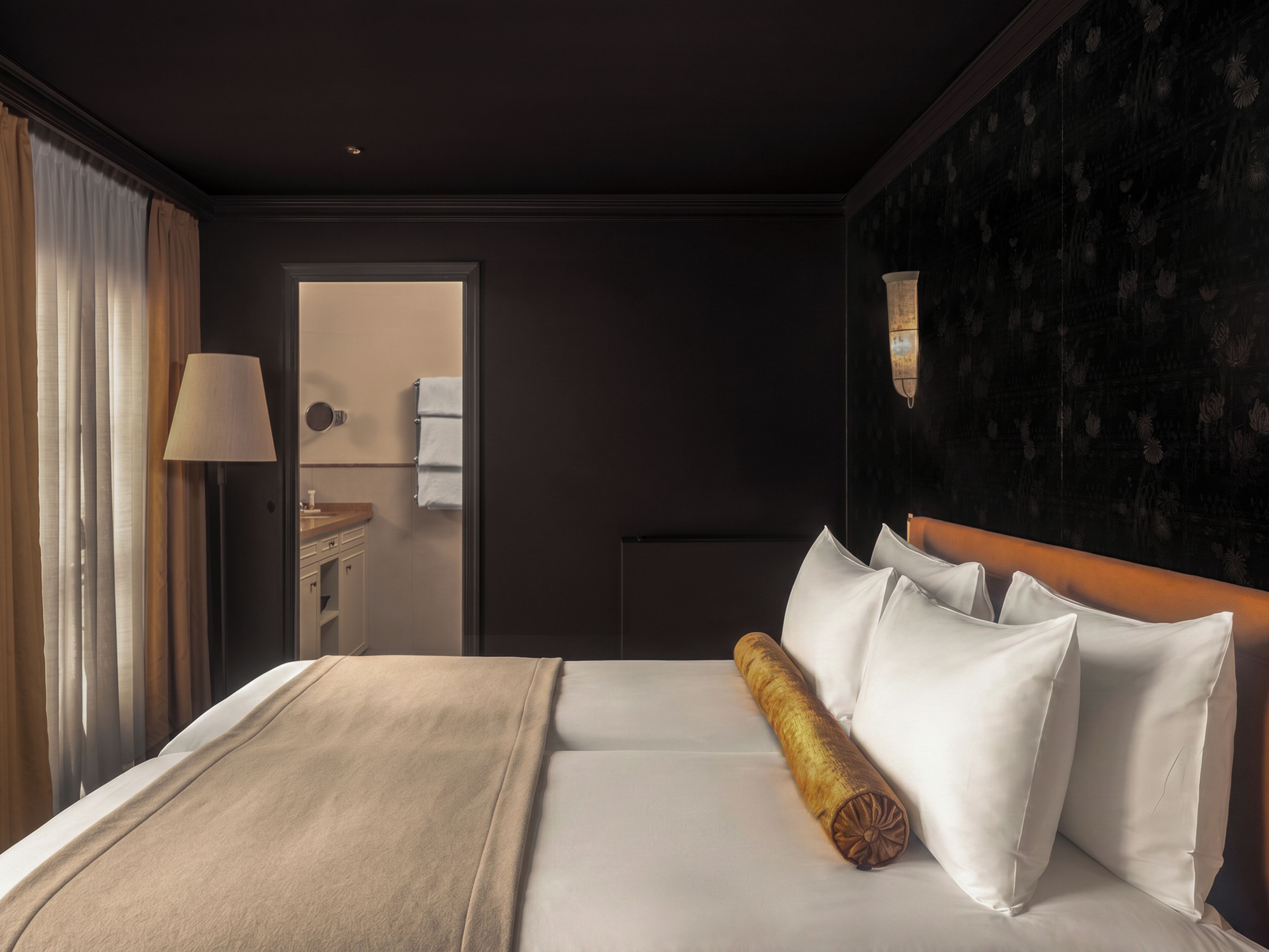 A hotel room with a bed, a nightstand, a lamp, a bathroom visible through an open door, and a dark wall with a floral pattern.