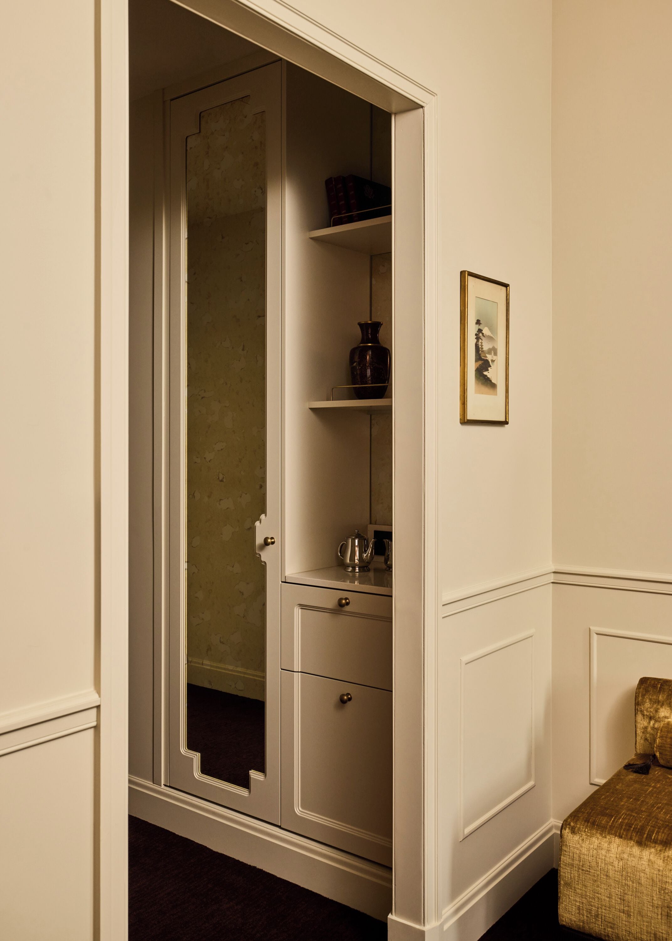 A room with a white wardrobe, a small shelf with books, a framed picture, and a gold-colored chair.