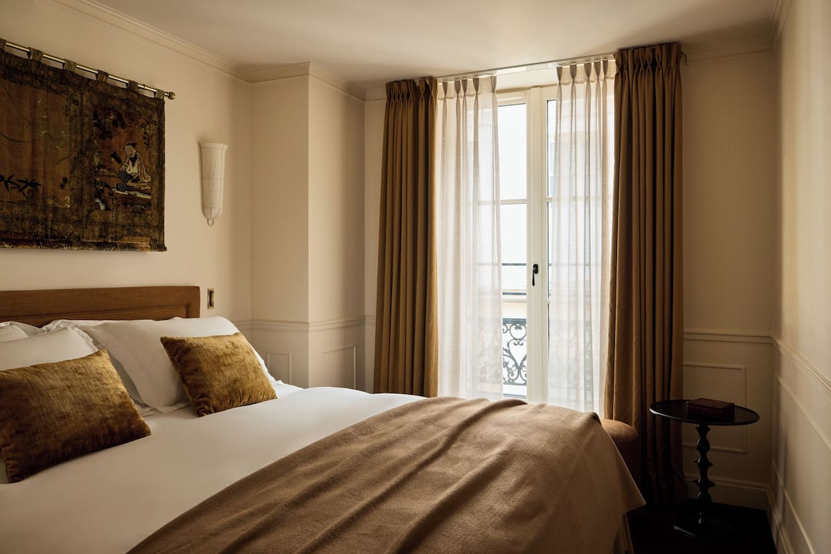 A neatly made bed with a brown blanket and pillows, a painting on the wall, and a window with curtains.