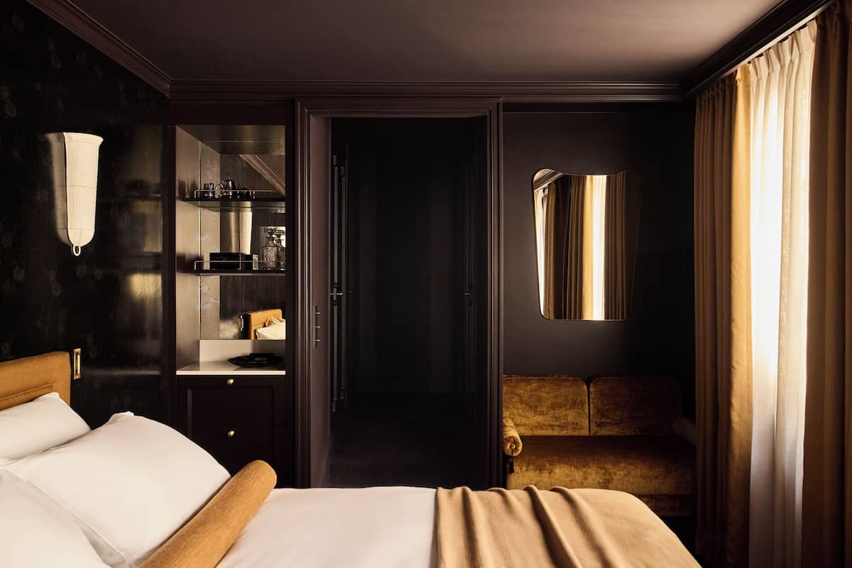 A hotel room with a bed, a vanity with a mirror, a small seating area, and a view of the city through the window.