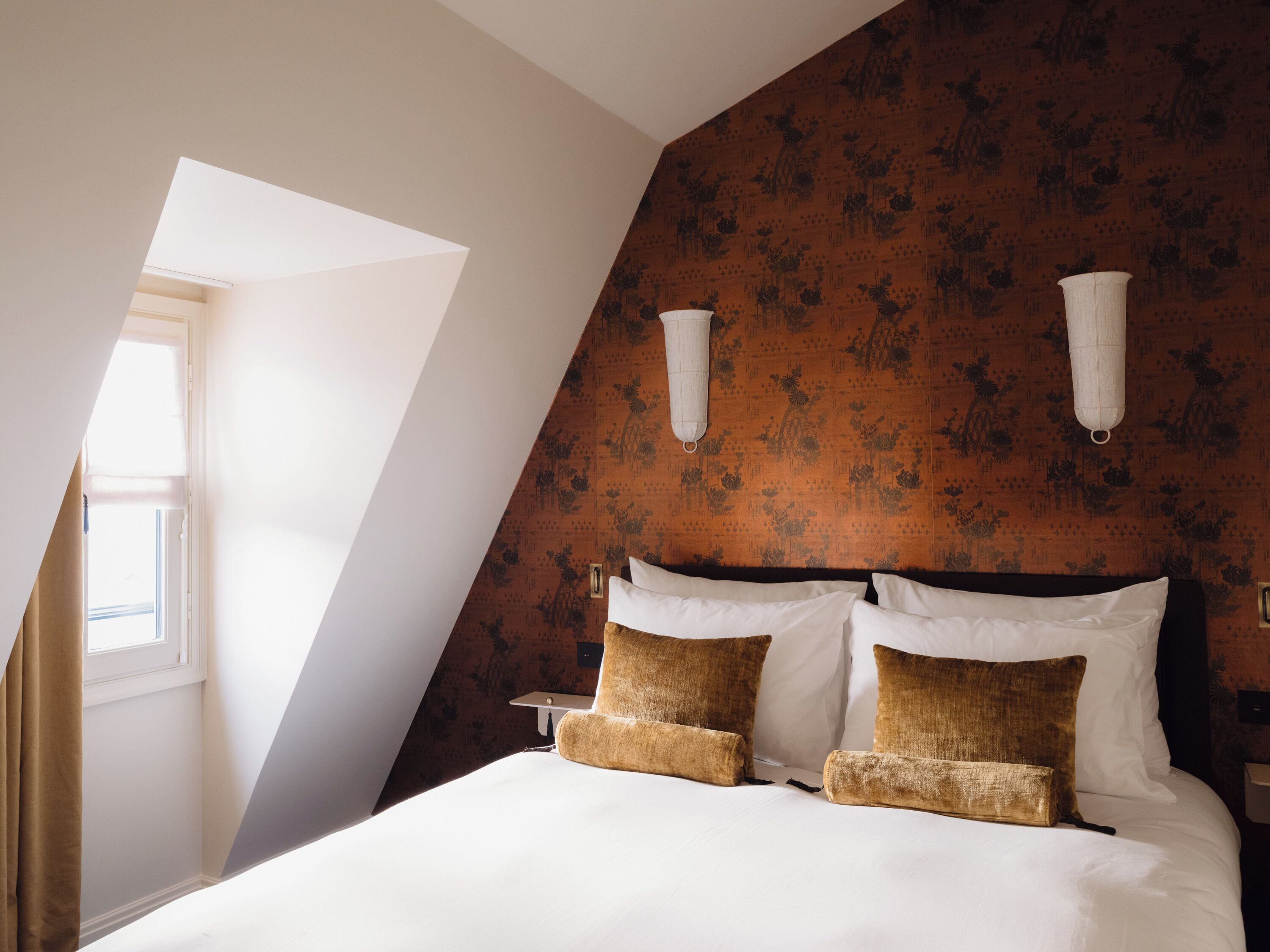 Un dormitorio con techo inclinado, cama con ropa de cama blanca y marrón, dos lámparas fijadas a la pared y una ventana con cortinas.