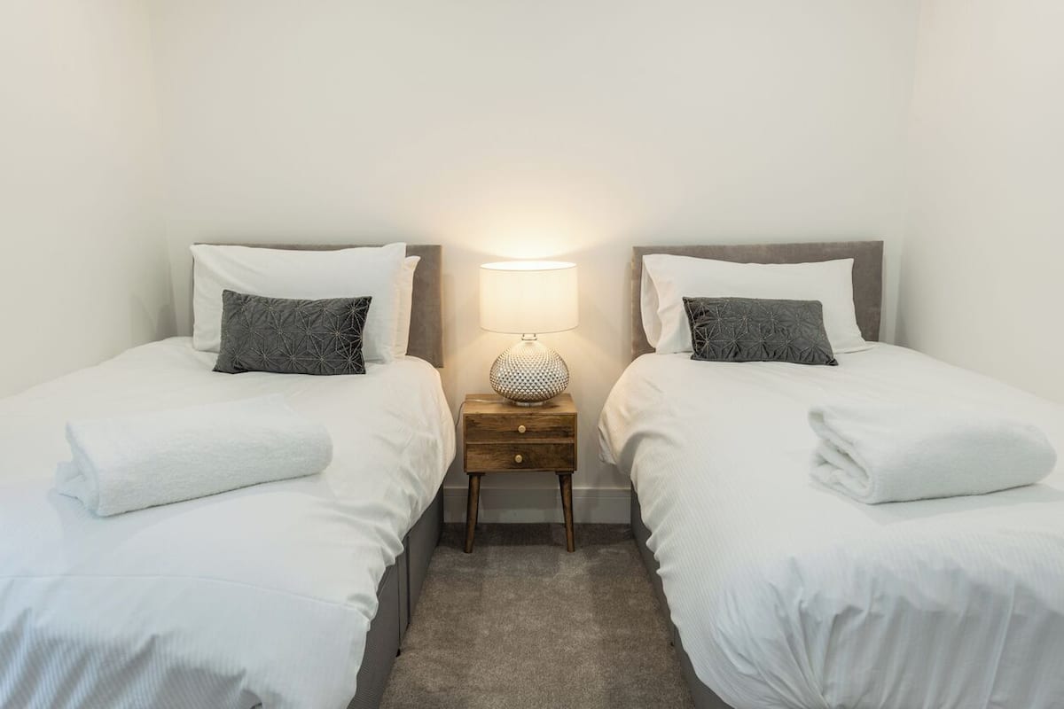 A hotel room with two single beds, each with a white duvet and a gray pillow. There is a small wooden nightstand with a lamp between the beds.
