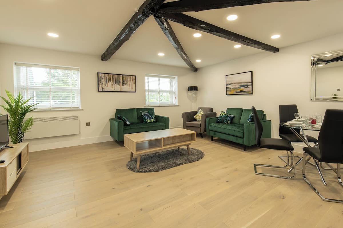 A modern living room with a wooden floor, green sofas, a coffee table, a dining table with chairs, and a large mirror.
