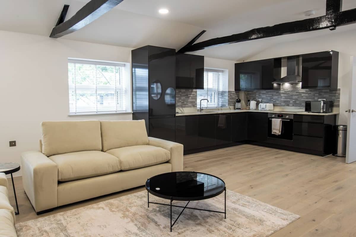 A modern living room with a beige sofa, a black coffee table, and a kitchen area with dark cabinetry and a tiled backsplash.