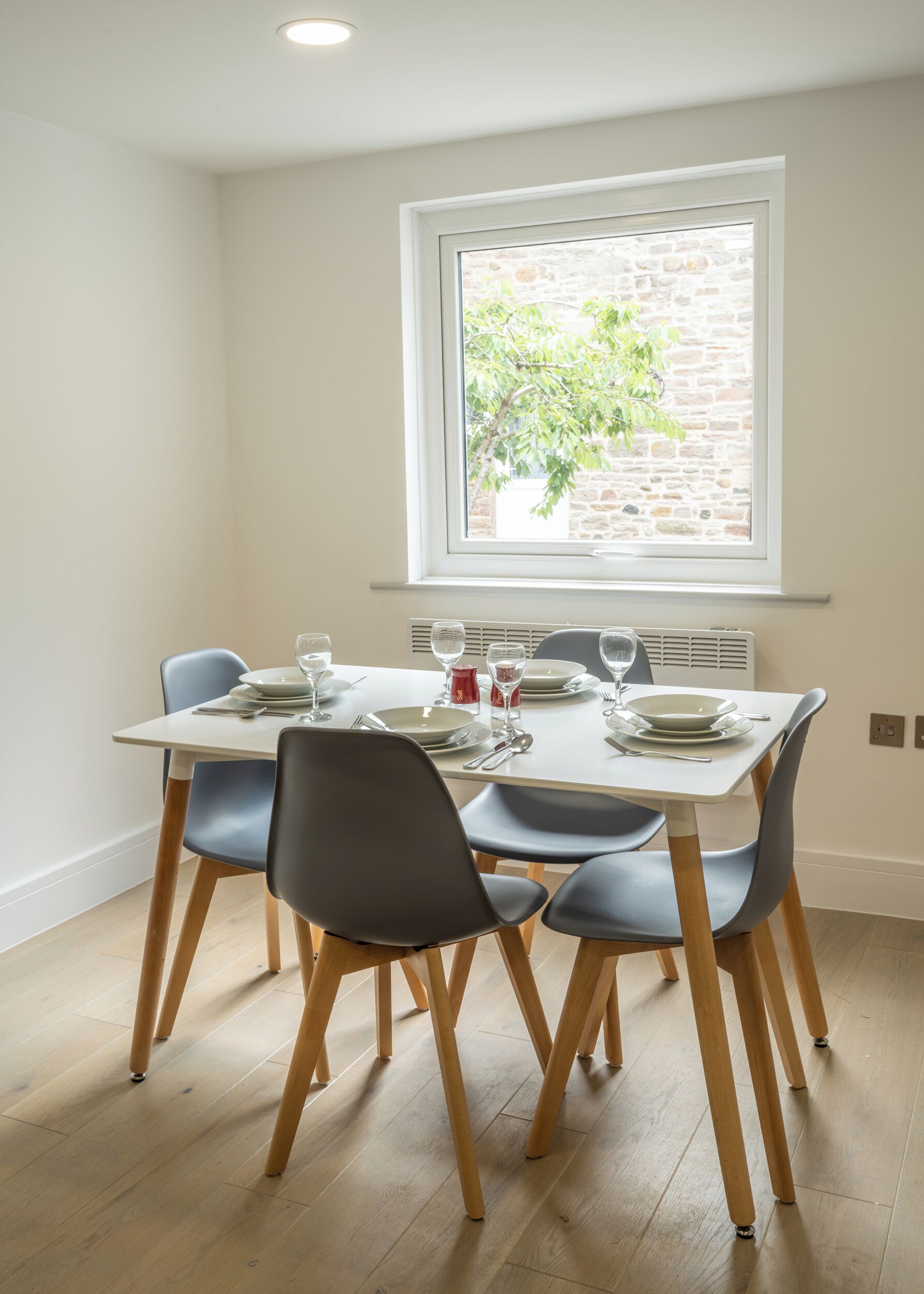 A dining area with a table set for four, four chairs, a window with a view of a brick wall and greenery, and a ceiling light.