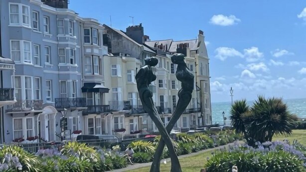 Un edificio frente al mar con una escultura en primer plano, cielo despejado y vista al océano.