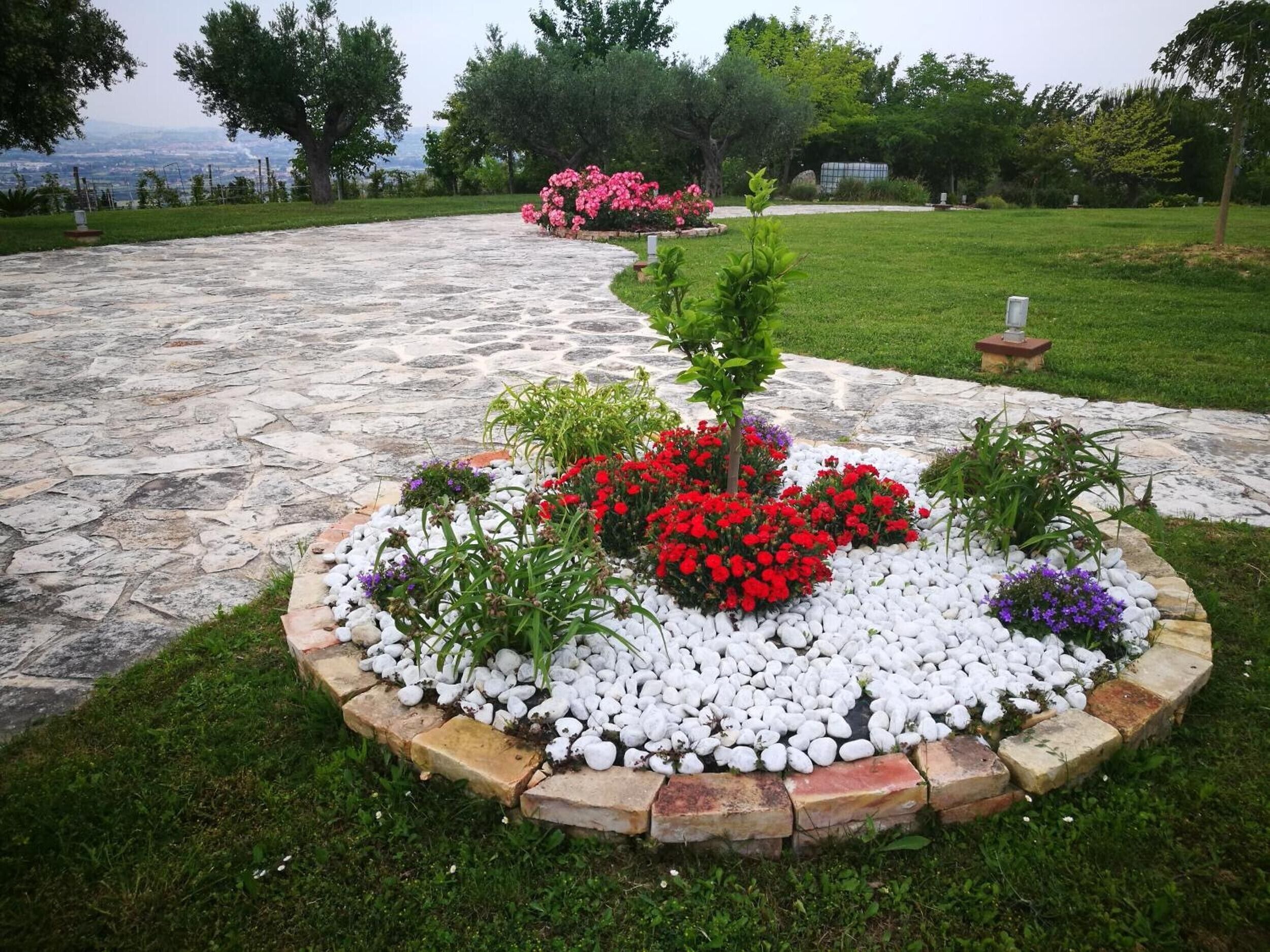 A stone pathway leading to a circular flower bed with red and purple flowers, surrounded by white pebbles and greenery.