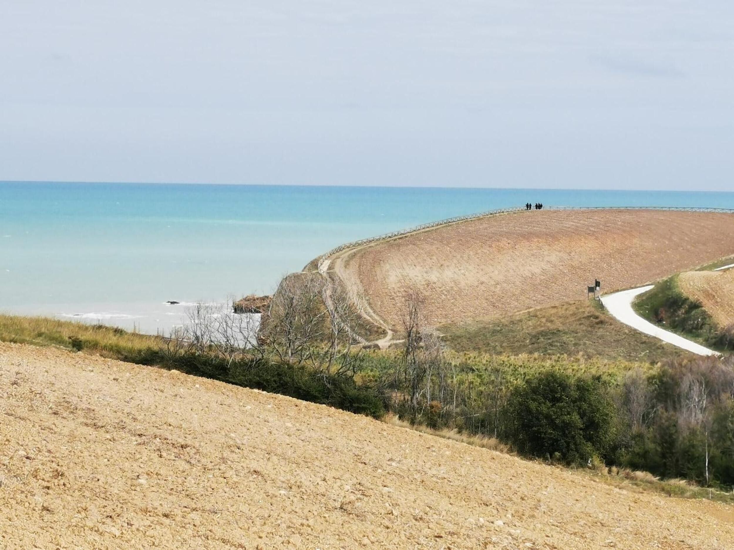 Un paisaje costero con un sendero de tierra que lleva a una playa, un área cercada y una vista despejada del mar.
