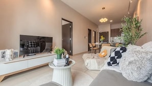 A modern living room with a flat-screen TV, a sofa, a coffee table, and a dining area in the background. - Bliss Residence Old Klang Road by FEEL HOME (Kuala Lumpur)
