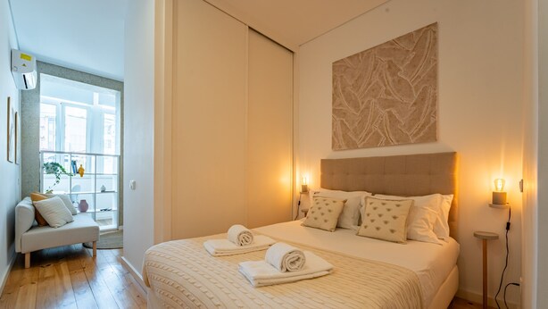 A bedroom with a large bed, two bedside tables with lamps, a sofa, and a window with a view of the city. - Nomad s Easy Stay - Duque Studio XIX (Porto)