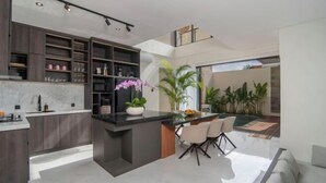 A modern kitchen with dark wood cabinets, a marble countertop, and a central island with chairs. There is a dining area with a pool visible through the glass wall. - Nidya Villa Canggu (Bali)