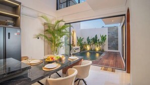A modern kitchen with a marble countertop, a dining area with two chairs, and a view of a swimming pool and outdoor plants. - Nidya Villa Canggu (Bali)