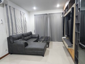 A modern living room with a sectional sofa, a coffee table, a flat-screen TV, and a built-in shelving unit. - Spacious Entire House With Shared Fitness Room and Shared Indoor Pool in Club (Pathum Thani)